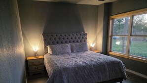 A bedroom with a tufted headboard, a bed with a grey quilt, two bedside lamps, and a window with a view of greenery. - Soothing 2-bedroom country Barndominium  in Beasley (Beasley)