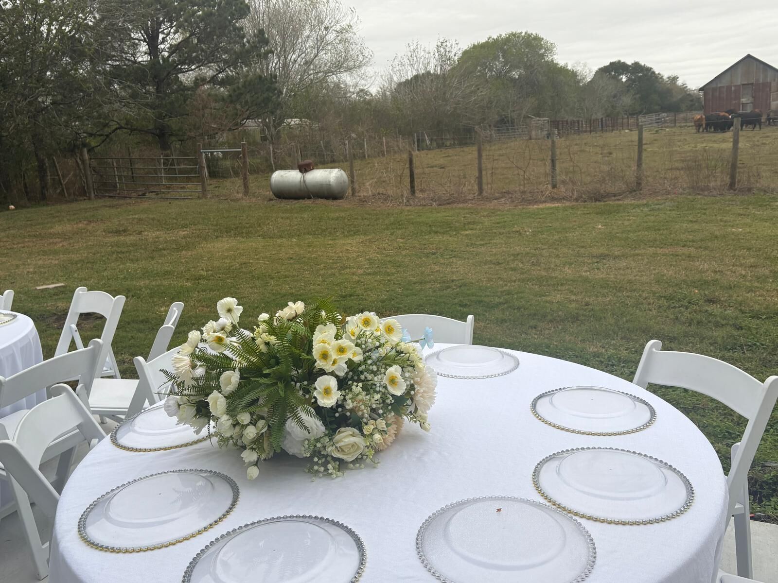 An outdoor dining area with round tables set for six, each with a white tablecloth and gold rimmed plates. A floral centerpiece adorns the center table.