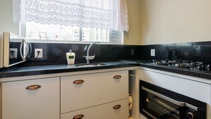 A kitchen with a black countertop, stainless steel appliances, a window with lace curtains, and a view of buildings outside.
