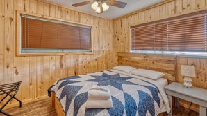 A wooden-paneled room with a bed, a ceiling fan, and a window with blinds.