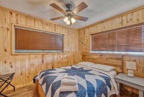 A wooden-paneled room with a bed, a ceiling fan, and a window with blinds.