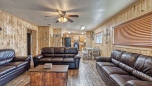 A wooden-paneled room with leather sofas, a coffee table, a ceiling fan, and a kitchen area in the background.