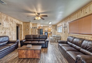 A wooden-paneled room with leather sofas, a coffee table, a ceiling fan, and a kitchen area in the background. - Bent River Cabin #2 @ Frio River Escape (Concan)