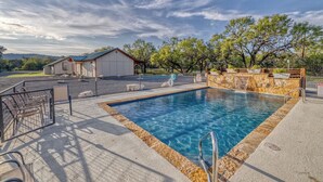 A rectangular swimming pool with a stone border, surrounded by a fence and a covered seating area with chairs. - Bent River Cabin #5 @ Frio River Escape (Concan)