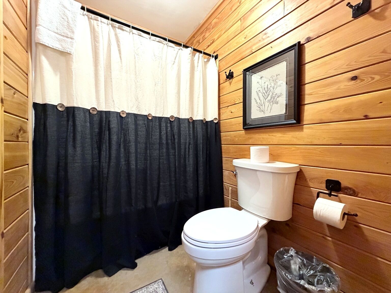 Un baño pequeño con paneles de madera, un inodoro, una cortina de ducha blanco y negro, e impresión botánica enmarcada en la pared.