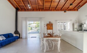 Een ruime woonkamer met houten plafond, een eettafel gedekt voor vier personen, een blauwe bank en een keuken met koelkast en fornuis.