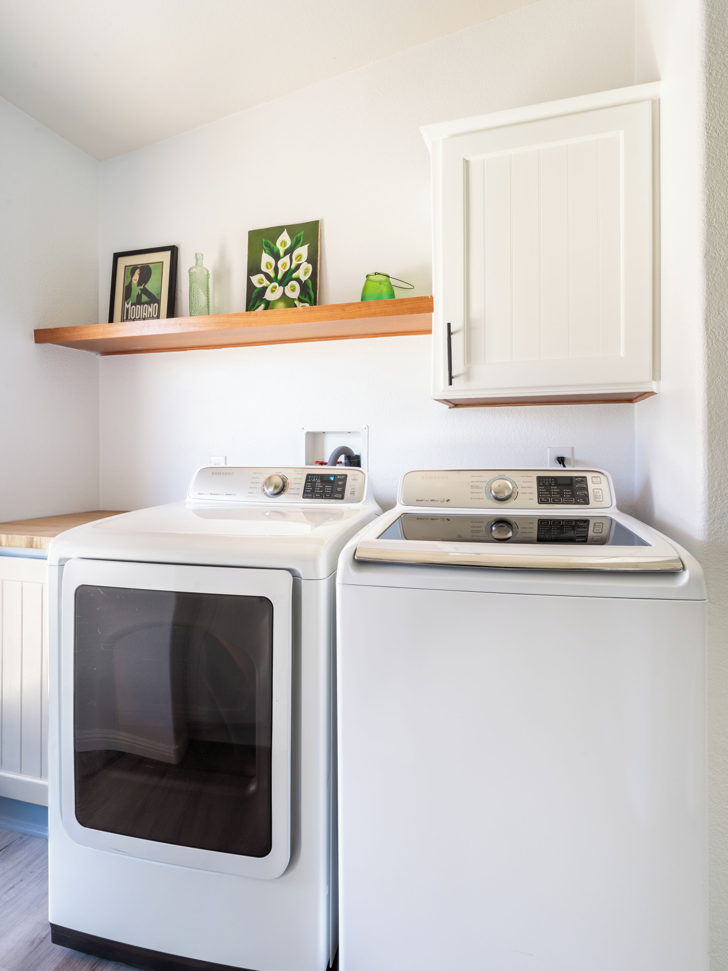 A laundry area with two front-loading washers, a wooden shelf with decorative items, and a white cabinet above.