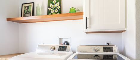 A laundry area with two front-loading washers, a wooden shelf with decorative items, and a white cabinet above.