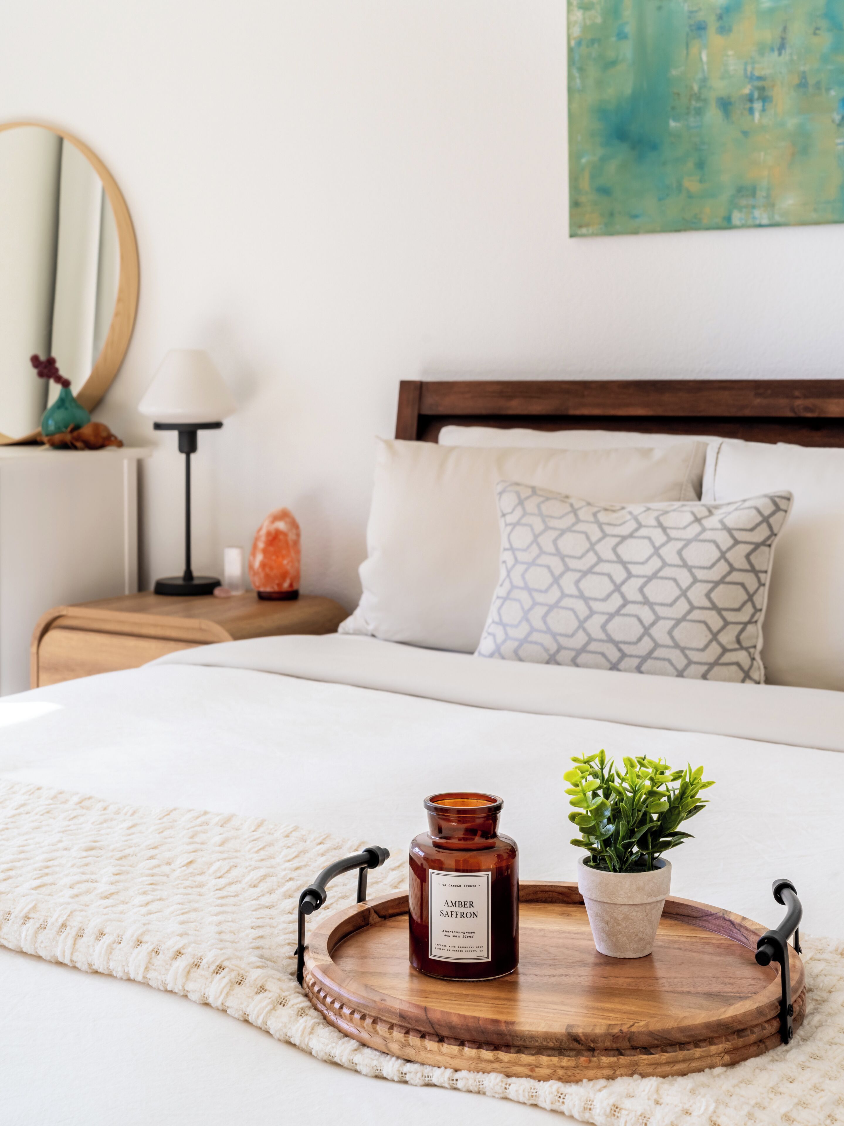 Een netjes opgemaakt bed met een houten dienblad waarop een kaars en een plant staan, een bedlamp en een decoratief object op het nachtkastje.