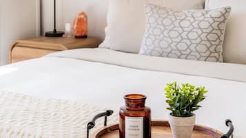 Een netjes opgemaakt bed met een houten dienblad waarop een kaars en een plant staan, een bedlamp en een decoratief object op het nachtkastje.