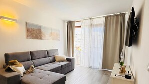 Una sala moderna con un sofá seccional gris, un pequeño otoman, una mesita auxiliar blanca con una botella y un control remoto, y un ventanal con cortinas translúcidas.