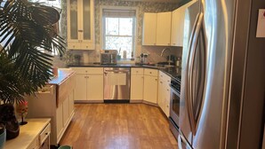 A modern kitchen with white cabinets, a stainless steel refrigerator, a dishwasher, and a window with blinds.