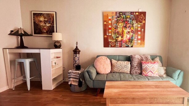 A cozy living room with a wooden coffee table, a green sofa with patterned cushions, a white desk with a lamp, and a colorful abstract painting on the wall.