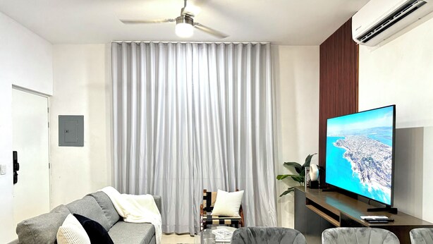 Uma sala de estar moderna com um sofá cinzento, uma mesa de centro de vidro, um ventilador de teto e uma TV de tela plana exibindo uma paisagem costeira.