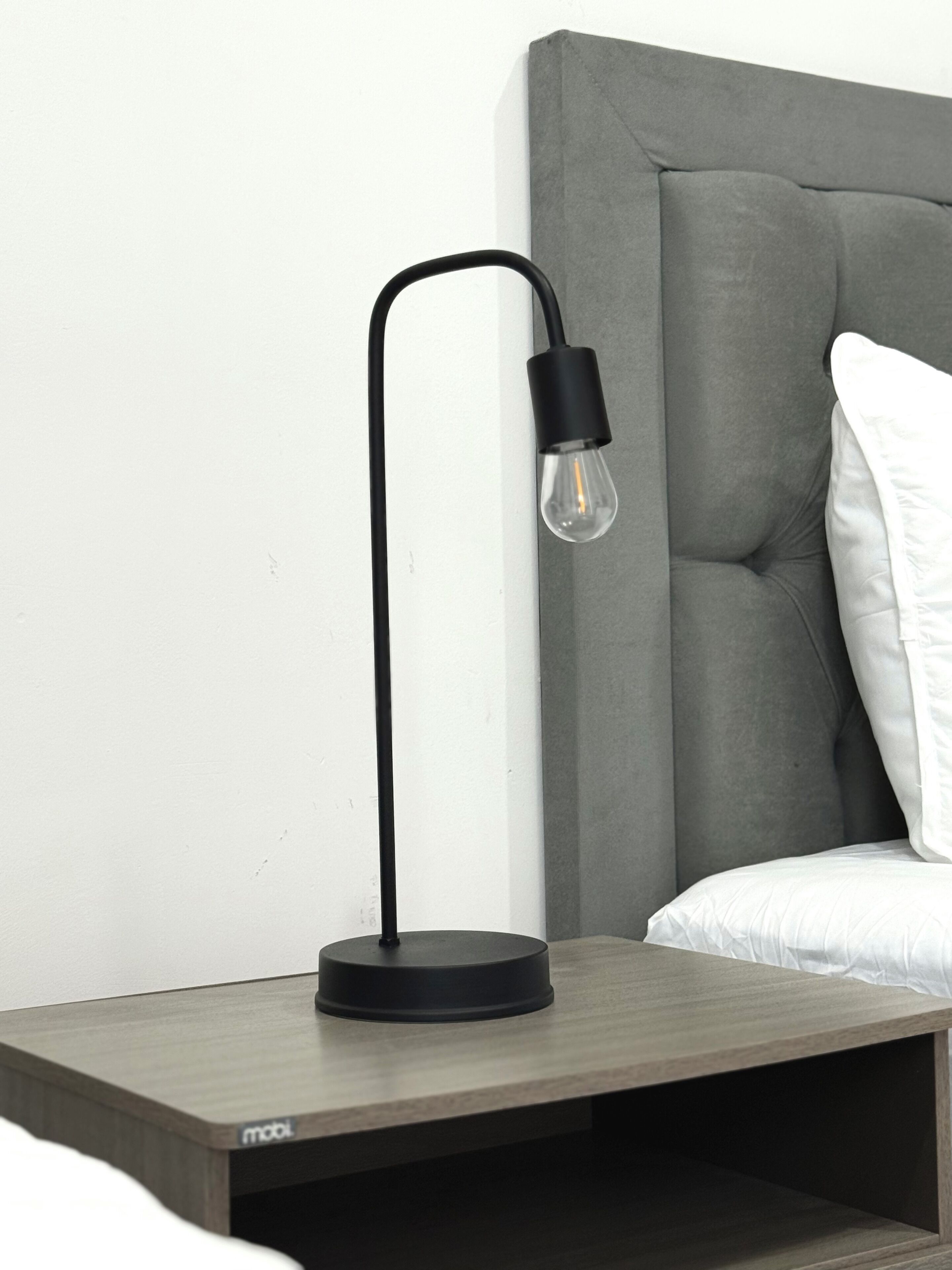 A modern bedside table with a black lamp, a grey upholstered headboard, and white bedding.