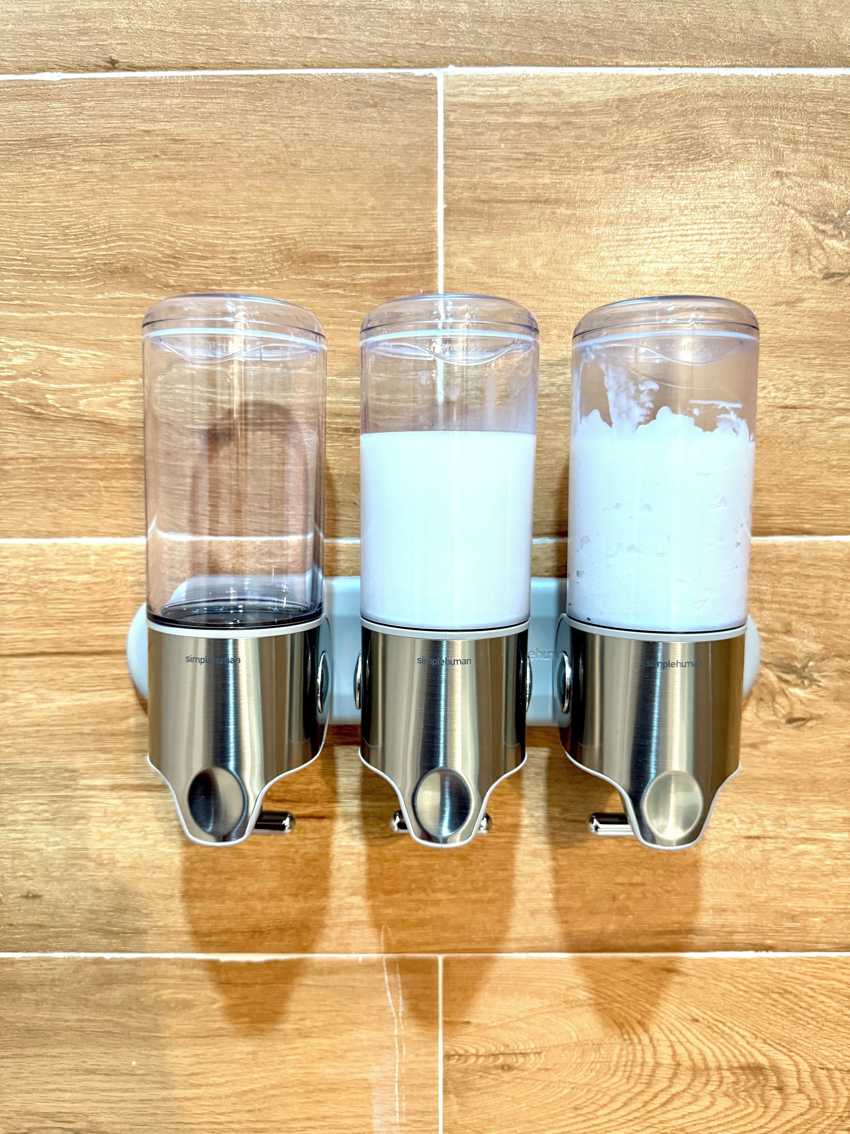 Trois distributeurs en acier inoxydable, dotés de plateaux supérieurs en verre transparent et remplis d’un liquide blanc, sont fixés à un mur en bois.