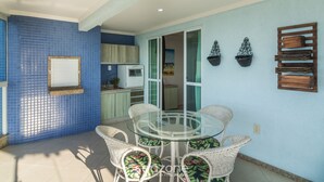 A dining area with a glass table and wicker chairs, a blue tiled wall, and a kitchen in the background. - Apto 3Qtos Vista Mar a 130m da Praia RDK0801 (Porto Belo)