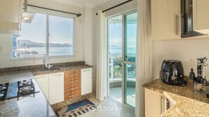 A modern kitchen with a gas stove, a coffee maker, and a view of the city through large windows. - Apto 3Qtos Vista Mar a 130m da Praia RDK0801 (Porto Belo)