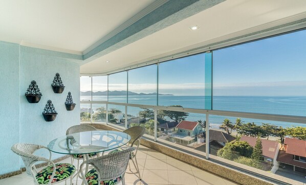 Un balcon avec une table en verre et des chaises en osier, offrant une vue sur la côte, avec des maisons et des montagnes en toile de fond.