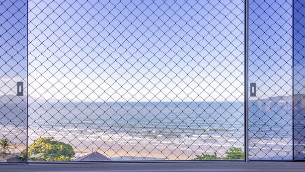 A view through a window with a chain-link fence, overlooking a beach with waves and buildings in the background. - Charming Sea View Apt in Porto Belo Rcs701 (Porto Belo)