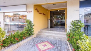 A hotel entrance with a yellow wall, a sign reading "Res. Costa do Saupe," and a number "2169." There are steps leading to the entrance, and a small garden area with red flowers and green plants. - Charming Sea View Apt in Porto Belo Rcs701 (Porto Belo)