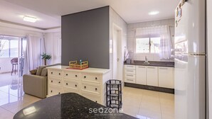 A modern kitchen with white cabinets, a black countertop, and a refrigerator. There is a dining area with a sofa and a plant. - Charming Sea View Apt in Porto Belo Rcs701 (Porto Belo)