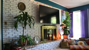 Una sala de estar con chimenea de ladrillo, un televisor de pantalla plana montado encima y una planta grande en maceta.