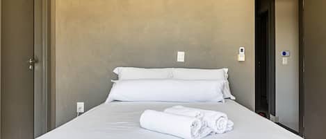 A neatly made bed with white linens and towels, a plain wall, and a door to the left.