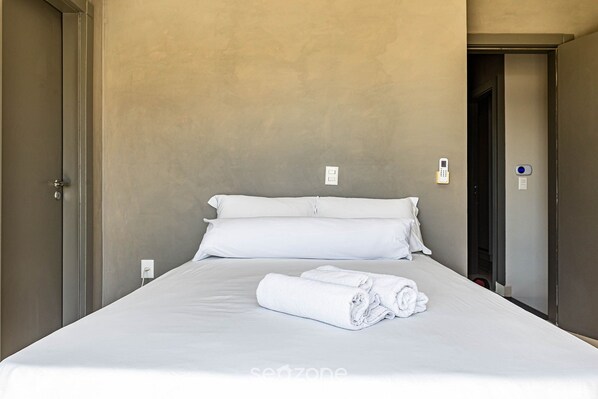 Un lit bien fait, avec du linge de lit et des serviettes blanches, un mur sobre et une porte sur la gauche.