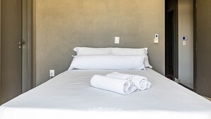 A neatly made bed with white linens and towels, a plain wall, and a door to the left. - Duplex 3Q pé na areia c/ vista deslumbrante RAT001 (Porto Belo)
