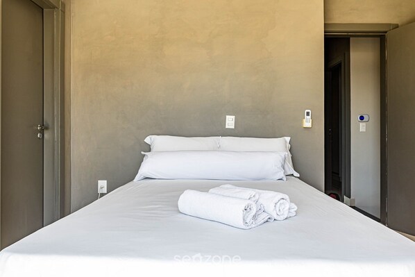 Una cama bien hecha con sábanas y toallas blancas, una pared lisa y una puerta a la izquierda.