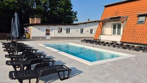 Ein Schwimmbecken mit Liegestühlen und Sonnenschirmen, ein Backsteinhaus mit einem Ziegeldach und eine Tür.