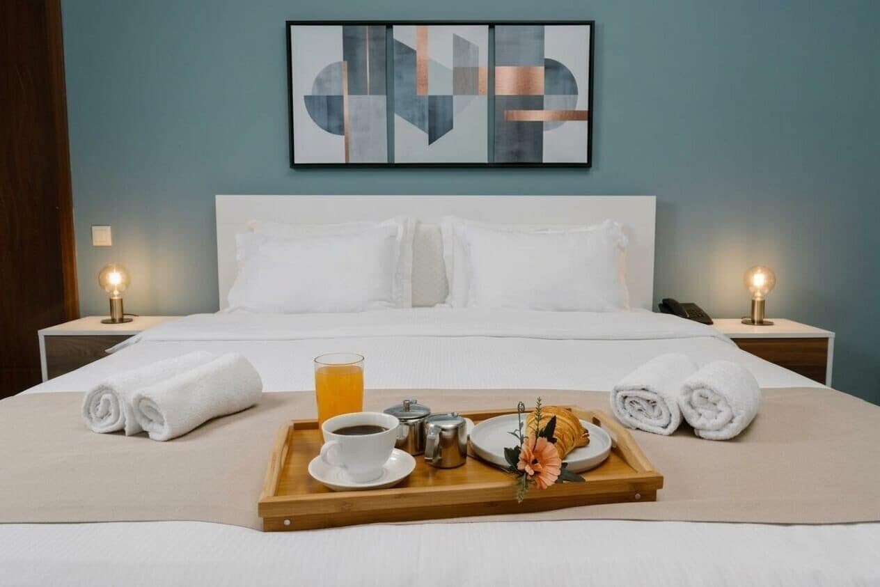 A hotel room with a neatly made bed, a tray with breakfast items, and a framed abstract artwork on the wall.