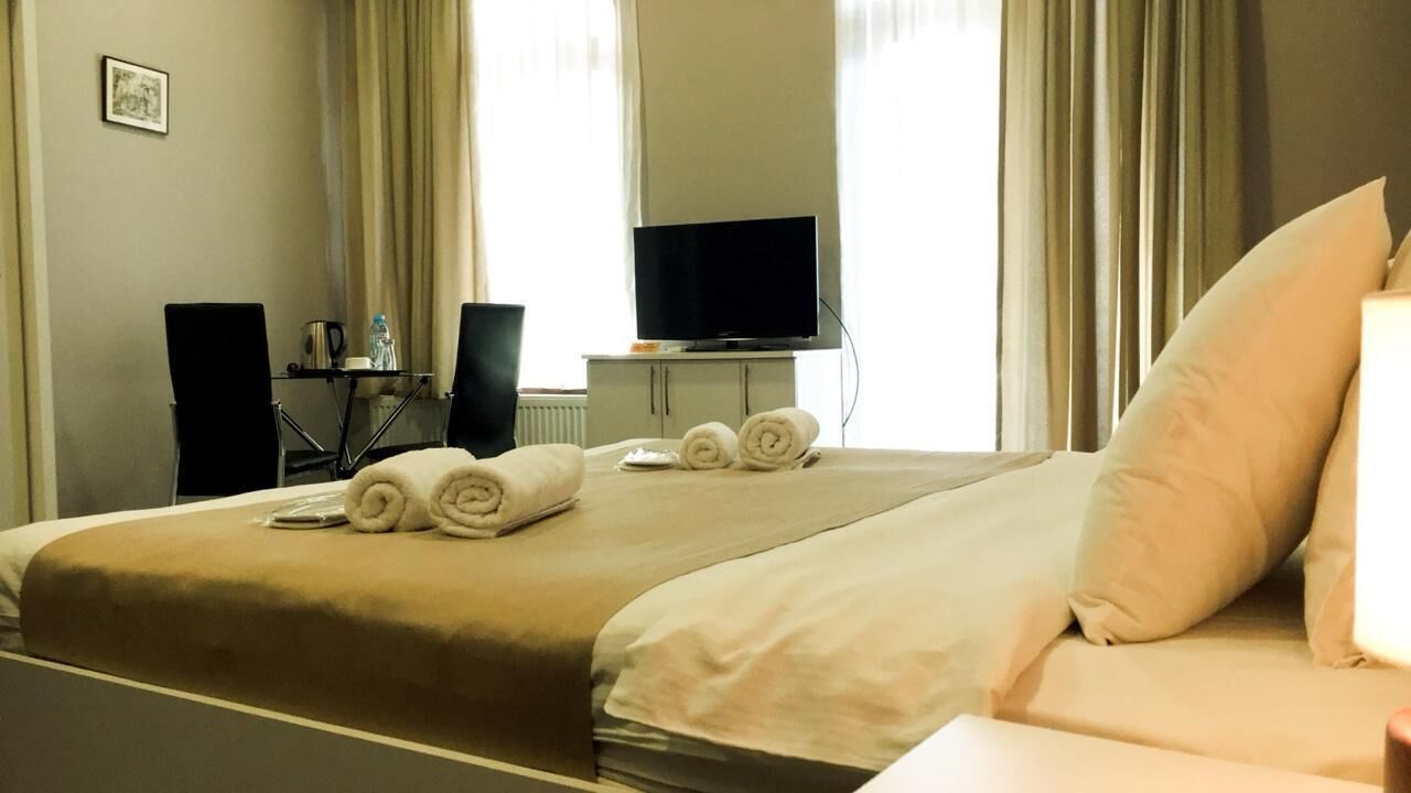 A hotel room with a bed, a television on a stand, a desk with a chair, and a window with curtains.