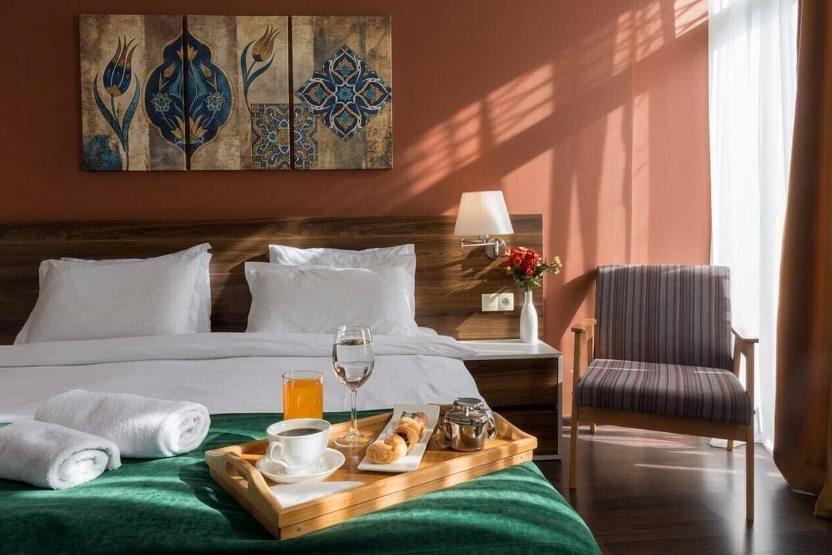 A hotel room with a bed, a tray with breakfast, a chair, and a wall with decorative art.