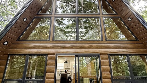 A modern cabin with a large glass front, wooden exterior, and a deck with outdoor seating.