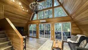 A spacious loft-style interior with a wooden staircase, a chandelier, and large windows offering views of the outdoors.