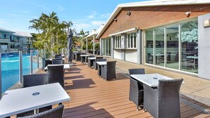 Una terraza exterior con mesas y sillas, una piscina y un edificio con ventanas amplias.