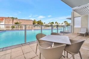 Zona piscina con tavolo e sedie, vista diretta sulla piscina e un edificio sullo sfondo con muro in mattoni e palme.