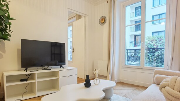 Un salon moderne avec un téléviseur à écran plat posé sur un meuble TV blanc, une table basse blanche, un canapé beige et une grande fenêtre donnant sur un immeuble et de la verdure.