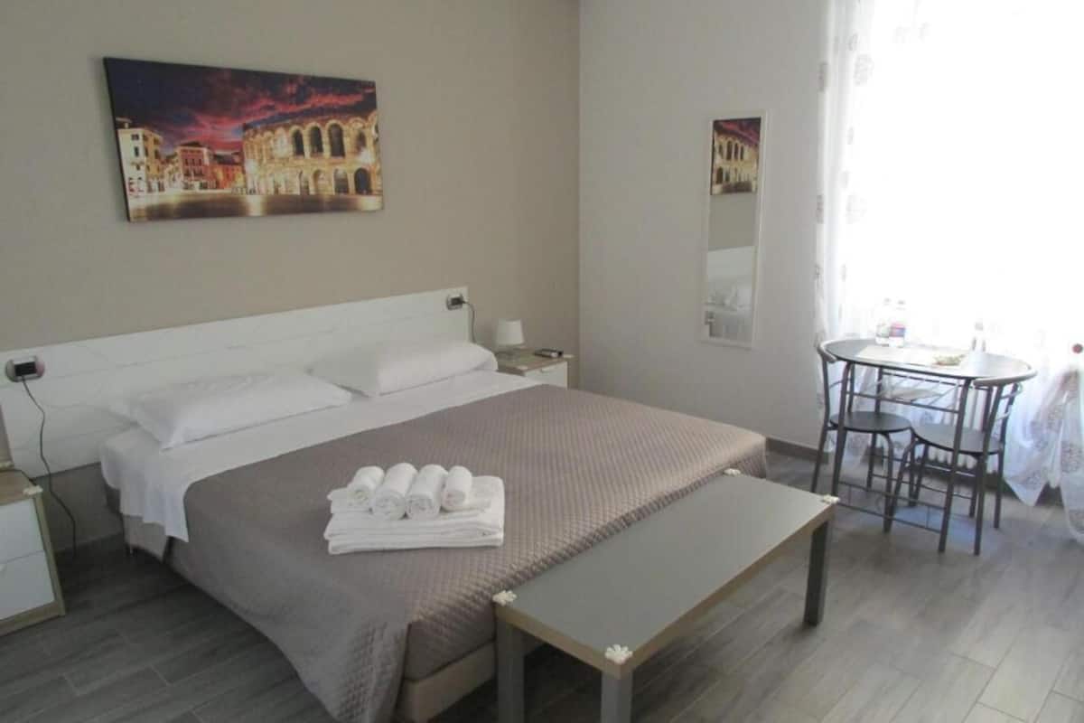 A hotel room with a bed, a bedside table, a chair, a small table, and a painting on the wall.