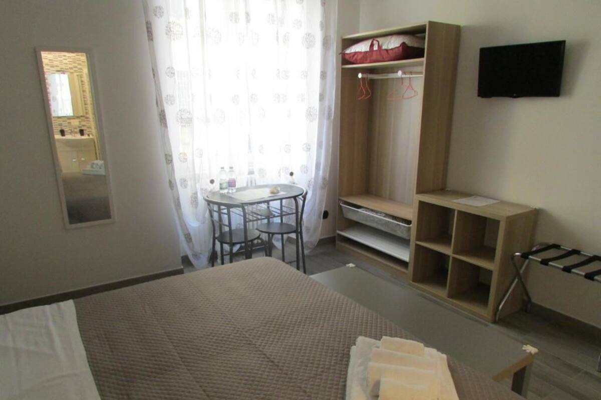 A hotel room with a bed, a small table with chairs, a mirror, a wardrobe, a TV, and a window with curtains.