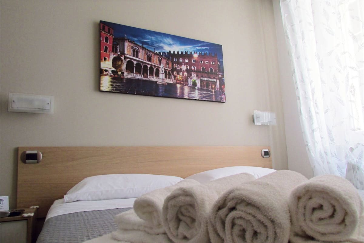 A hotel room with a bed, a painting of a bridge, and a window with curtains.