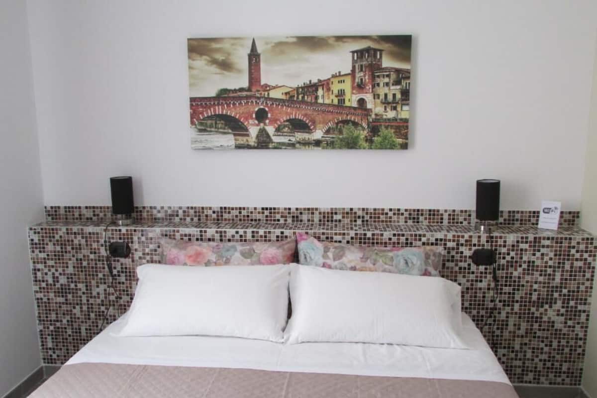 A hotel room with a bed, a wall-mounted painting of a bridge, and a tiled headboard.