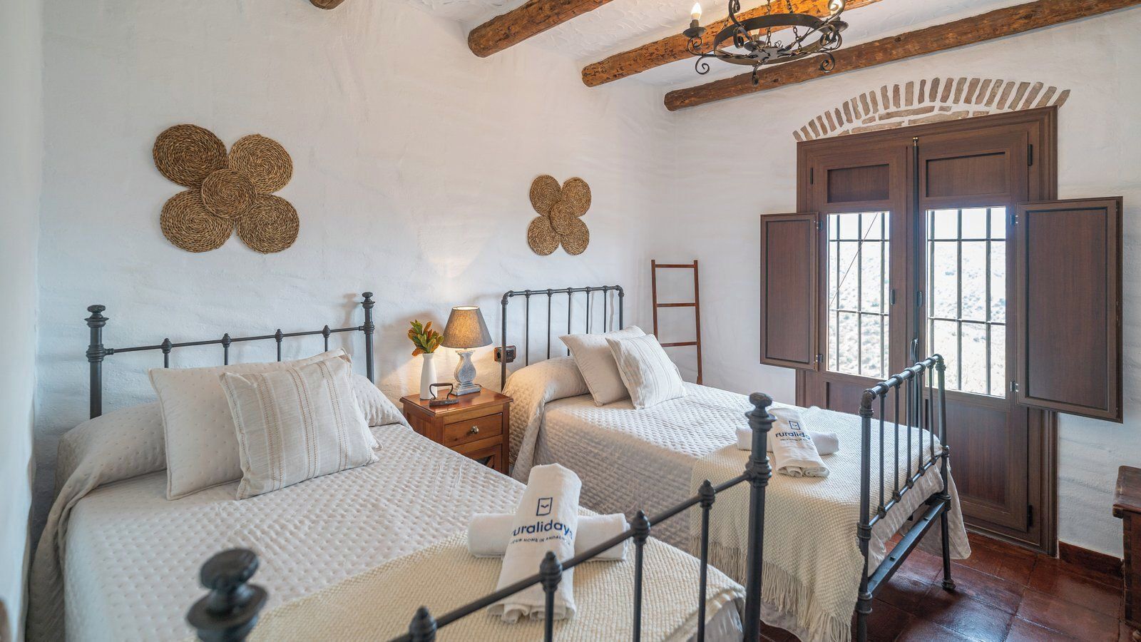 Un dormitorio con dos camas, una mesita de noche de madera con una lámpara y un ventanal con contraventanas de madera.