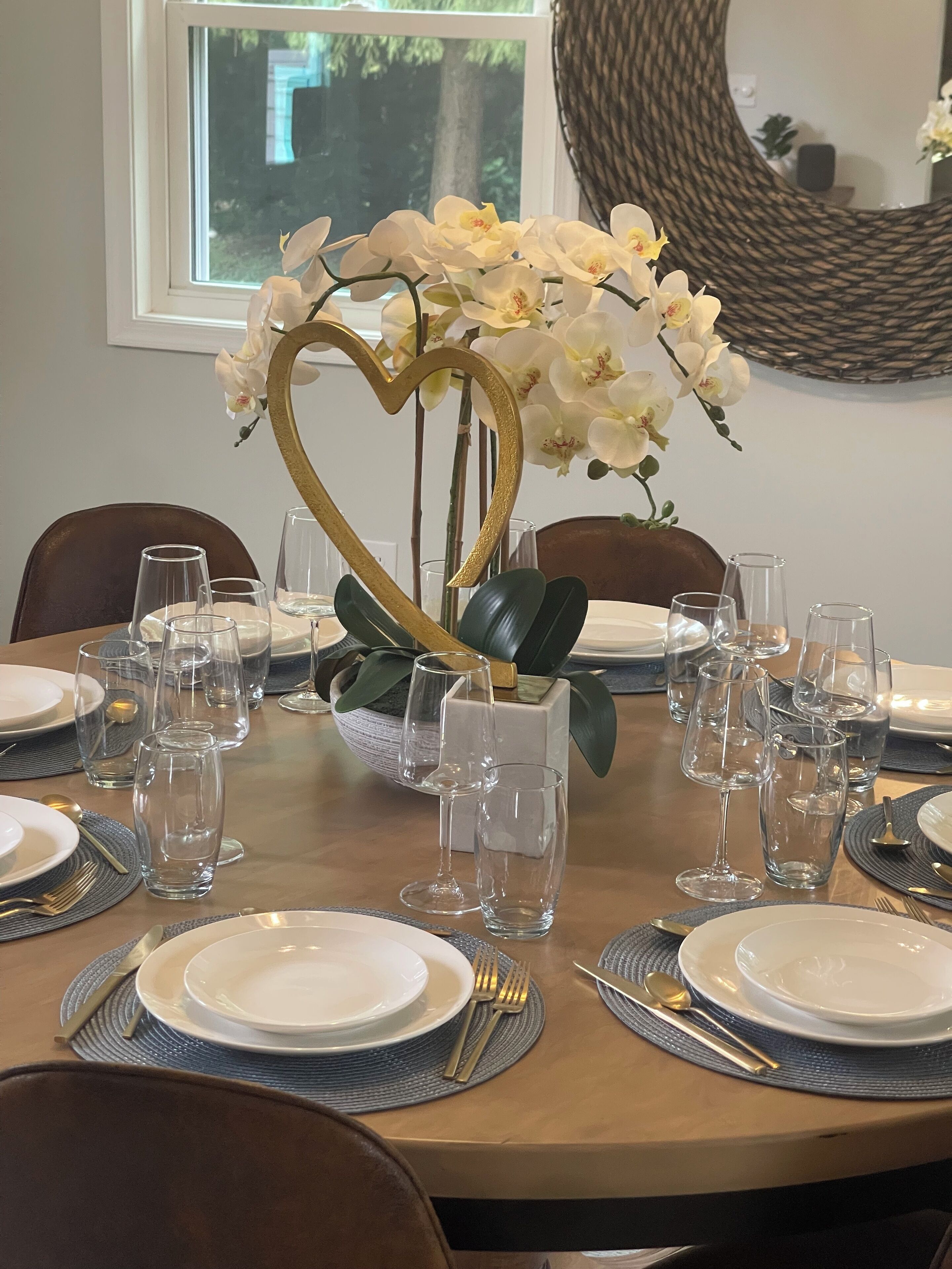 A dining table set with clear glassware, plates, and cutlery, centered with a golden heart-shaped orchid arrangement.