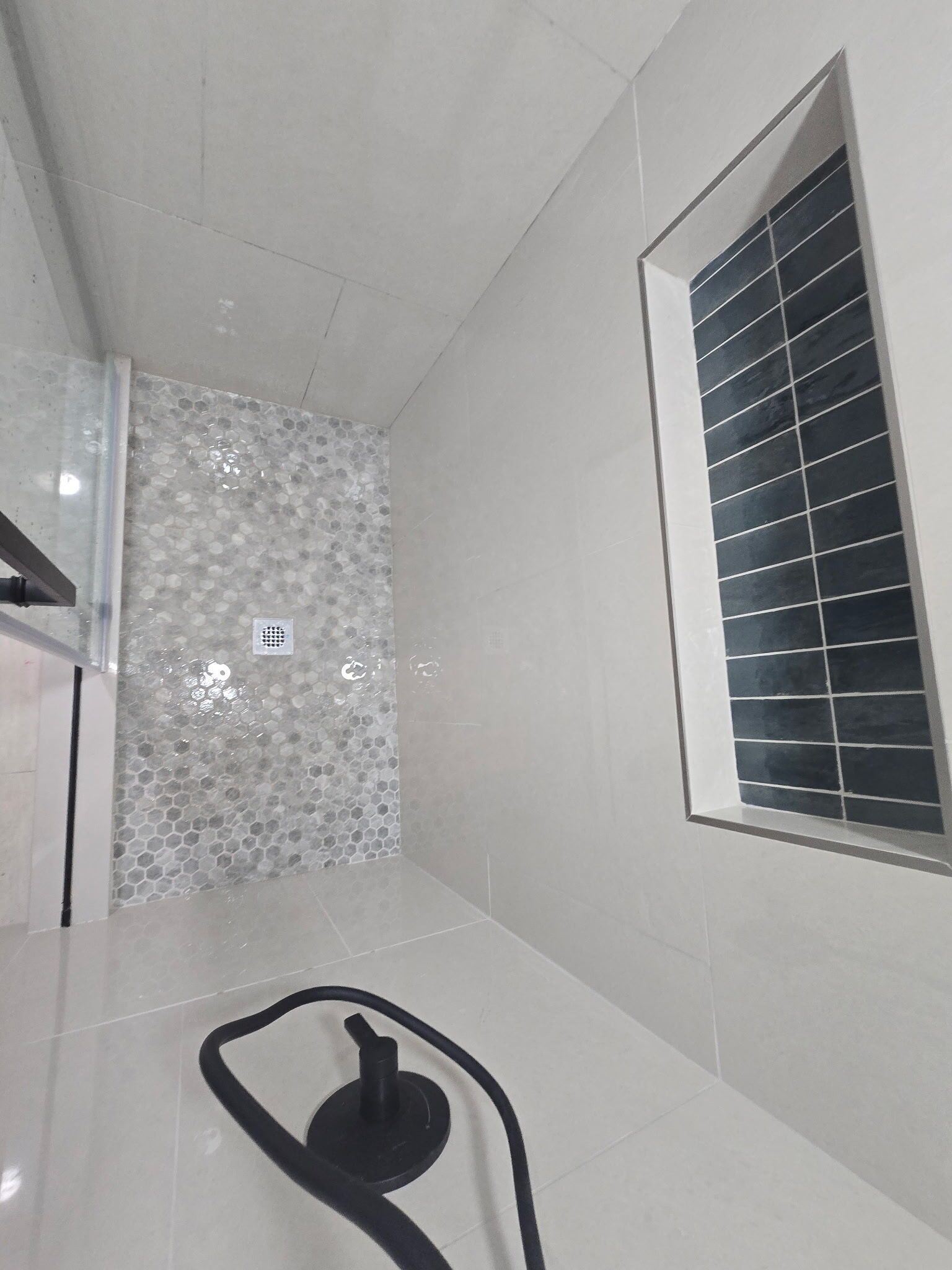 Un baño con piso de cerámica, zona de ducha con pared revestida de azulejos, ventana con persianas y un respiradero visible.
