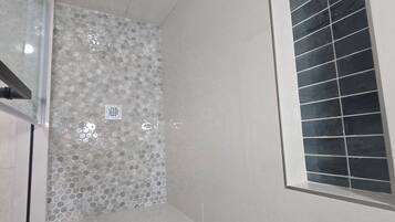 Un baño con piso de cerámica, zona de ducha con pared revestida de azulejos, ventana con persianas y un respiradero visible.
