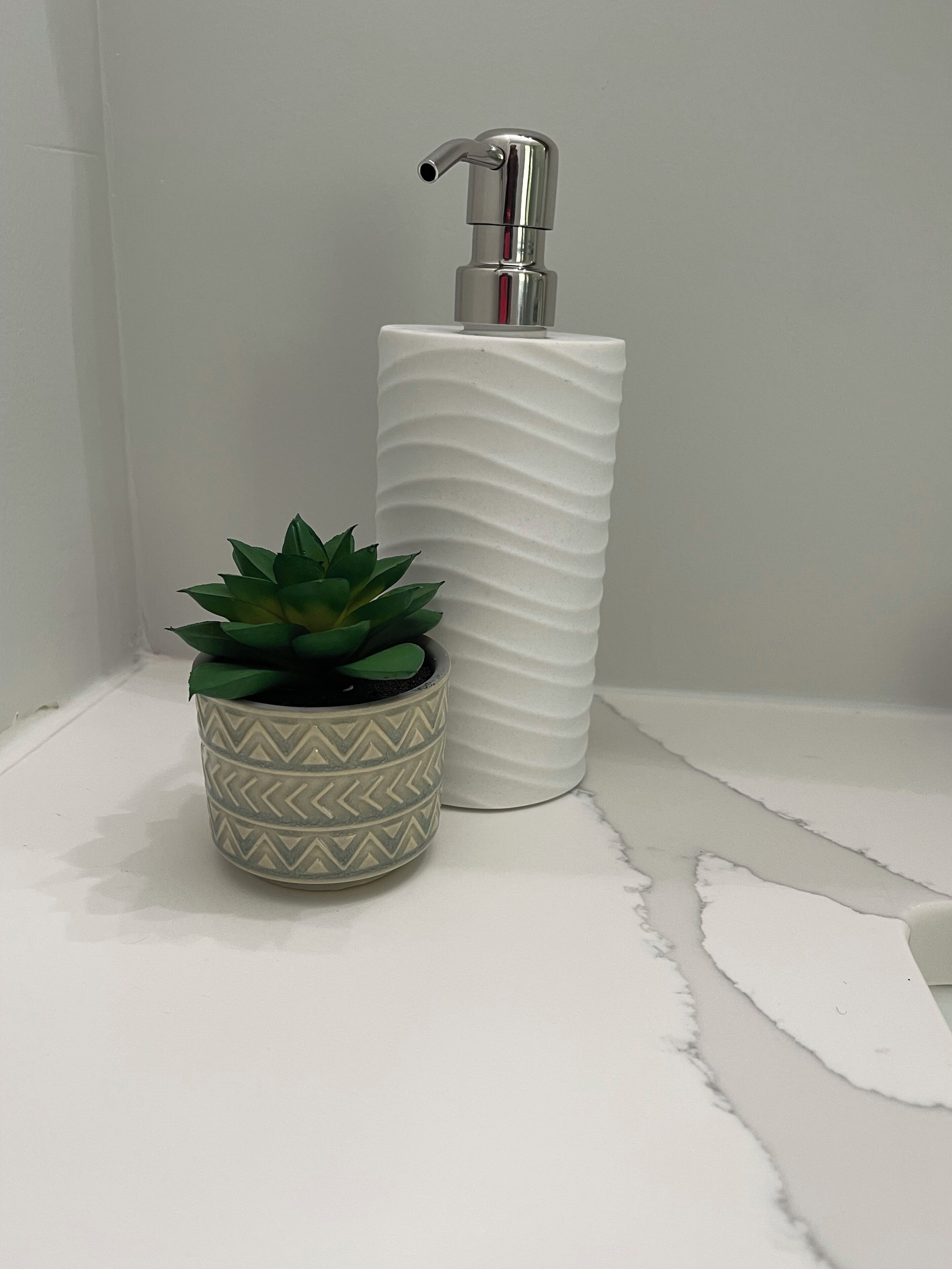 Un bagno con parete bianca strutturata, dispenser di sapone bianco e pianta in vaso con decorazioni.