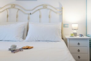 Un lit bien fait, avec du linge de lit blanc, une table de chevet avec une lampe, une tasse et une assiette contenant de la nourriture.